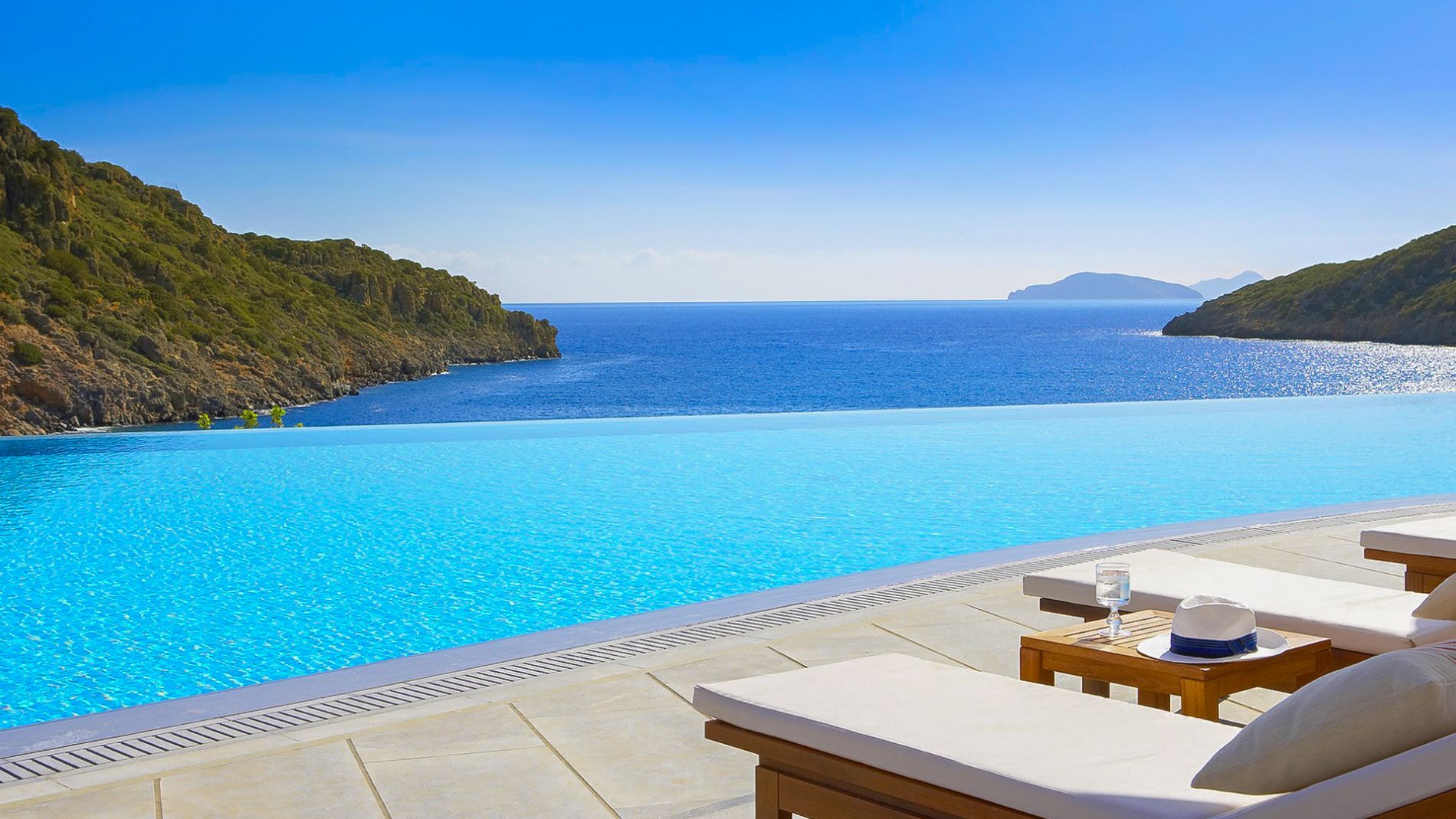 Infinity-Pool mit Meerblick vor Felsenküste und blauer See; Liegen mit Tisch, Hut und Glas am Pool.