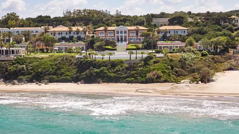Mandola Rosa Resort am Riviera Olympia: gepflegter Garten, Palmen, weiße Gebäudefronten am Hang über Strand und türkisfarbenem Meer.