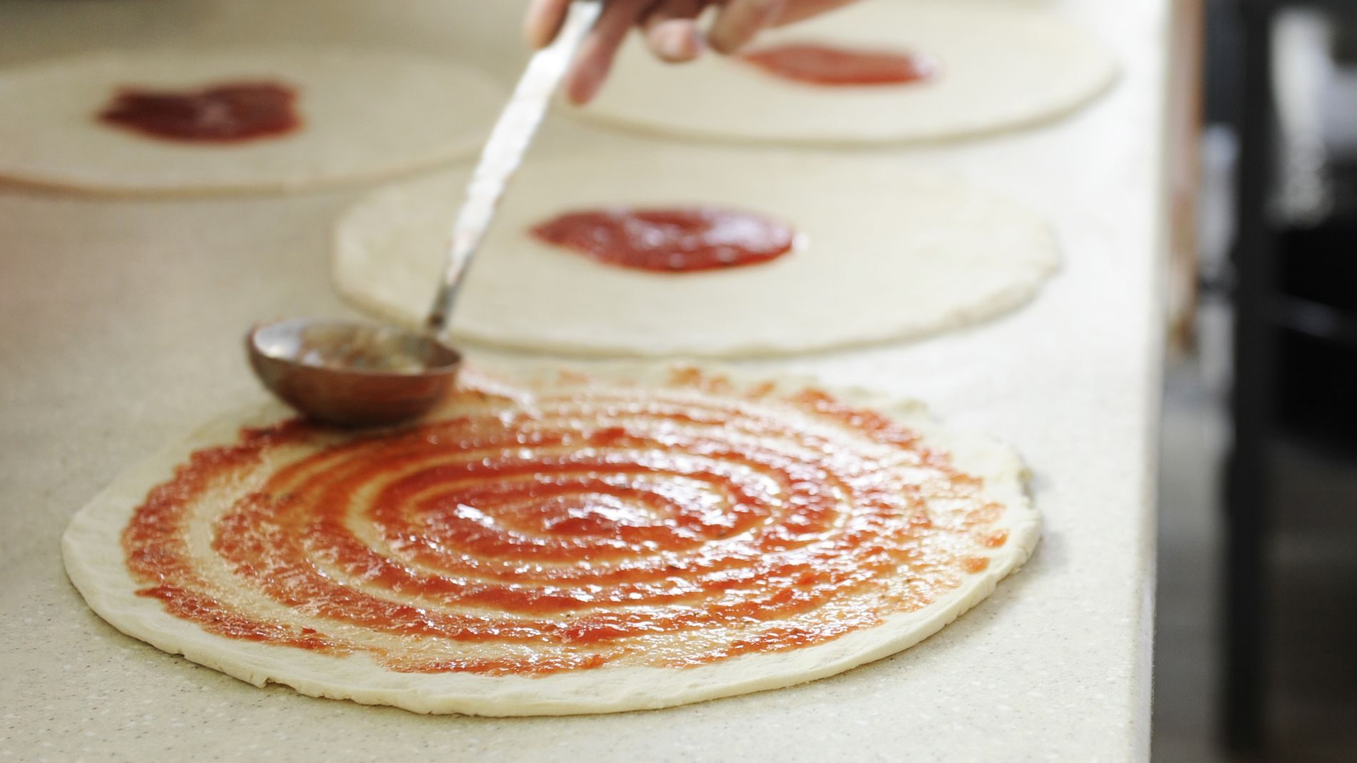 Küchenarbeit: Pizzateig liegt ausgerollt, rote Tomatensauce wird kreisförmig mit Löffel aufgetragen.
