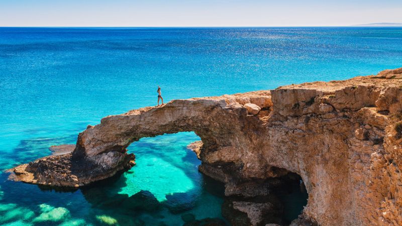 Frau steht auf einem Felsbogen über türkisblauem Meer; natürliche Steinbrücke vor der Zyprenküste bei Ayia Napa. Frau steht auf einem Felsbogen über türkisblauem Meer; natürliche Steinbrücke vor der Zyprenküste bei Ayia Napa.