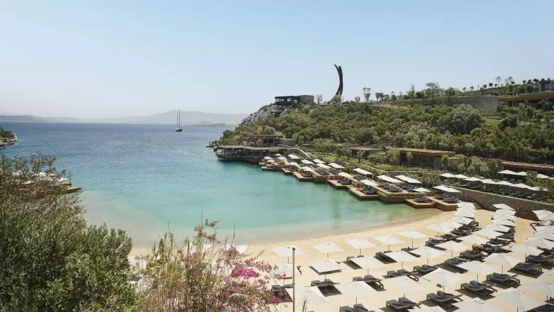 Strand von Maxx Royal Bodrum: turquoise Wasser, Sandstrand mit Liegen und Sonnenschirmen, Terrassen am Ufer, grüne Anhöhe mit Skulptur.