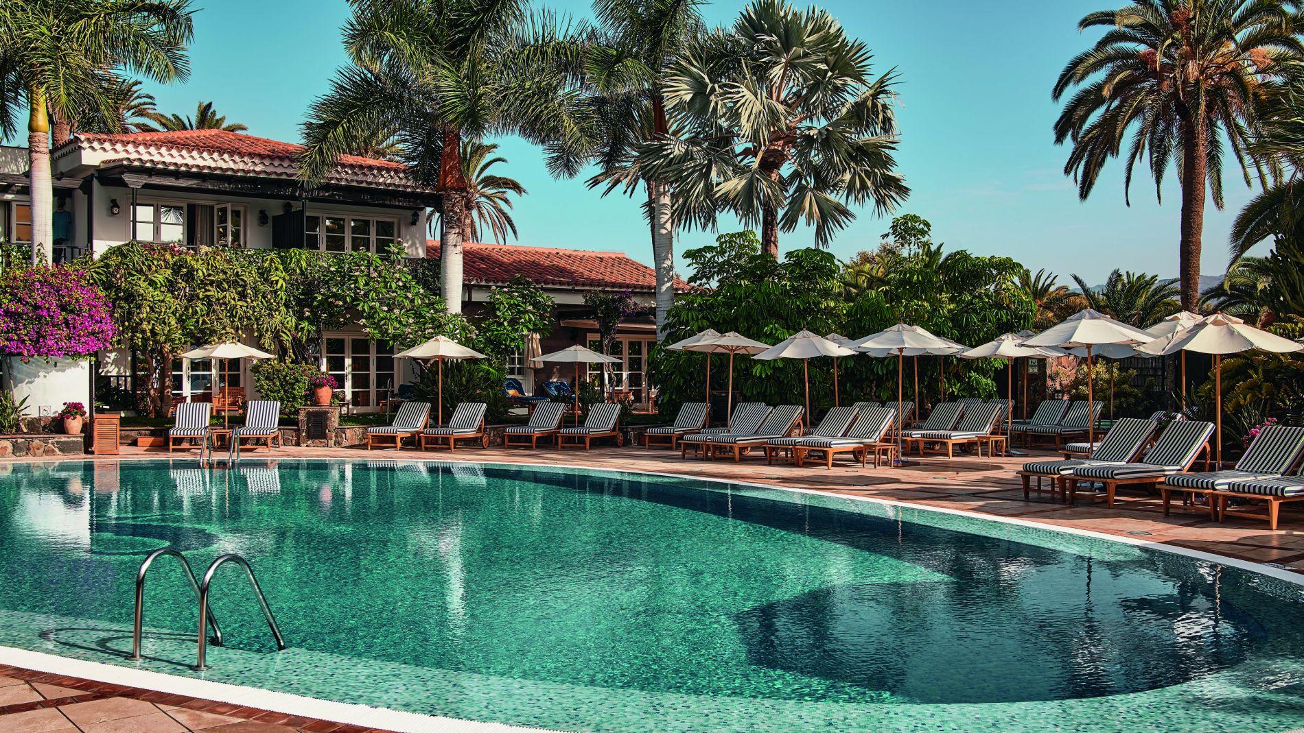 Außenpool mit türkisfarbenem Wasser, Liegen unter Schirmen, Palmen und Resort-Gebäude im Hintergrund