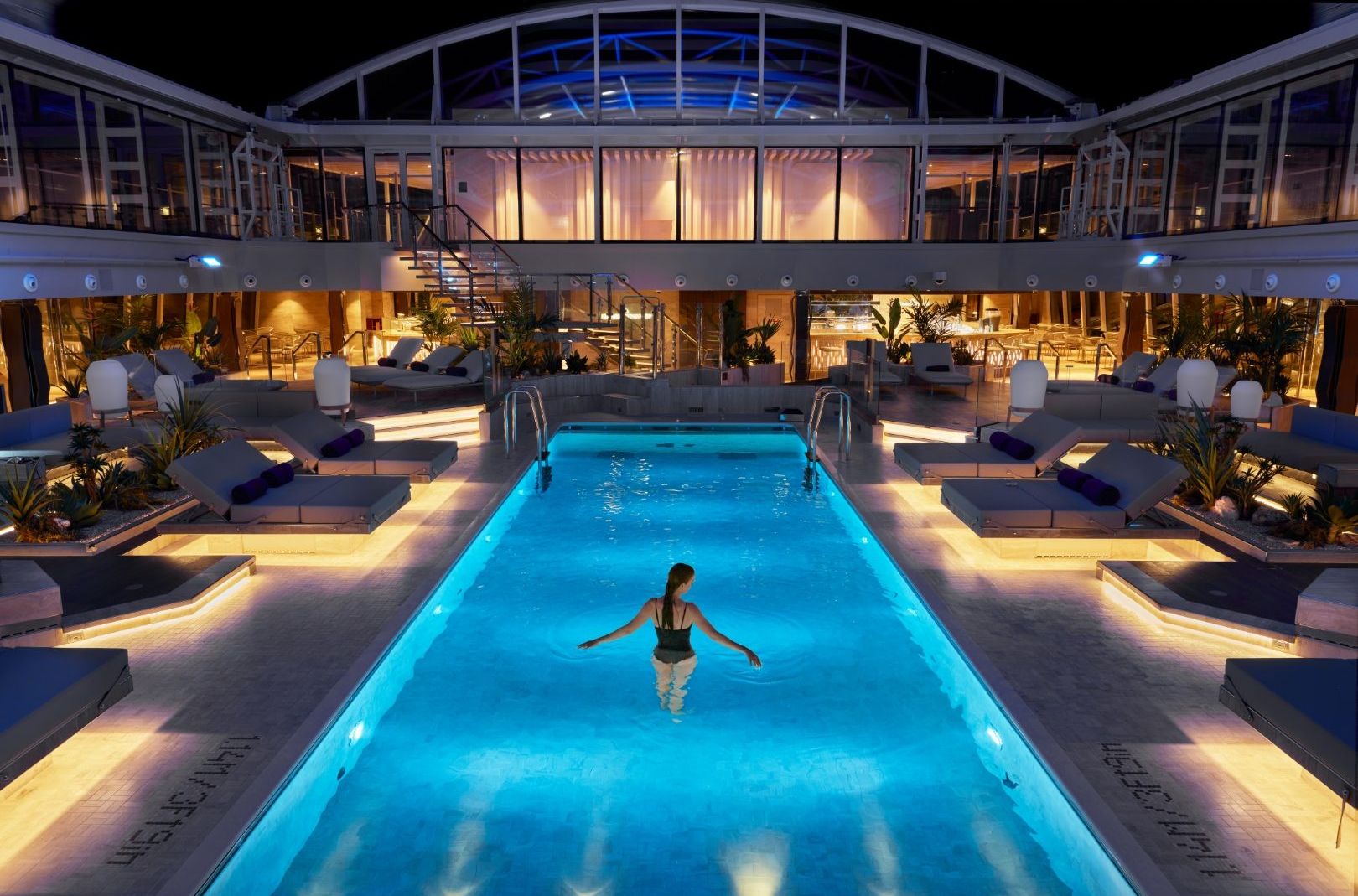 Innenpoolbereich eines Explora Journeys Schiffs, Abendbeleuchtung, schwimmende Person, umliegende Liegen und Pflanzen.