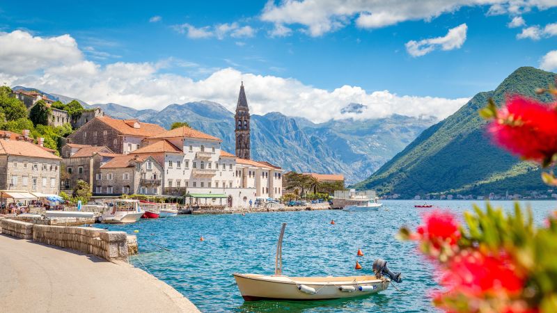 Historische Stadt Perast am Bay of Kotor, Montenegro; Kirchturm, Steinbauten, Boot am klaren Wasser, umliegende Berge.