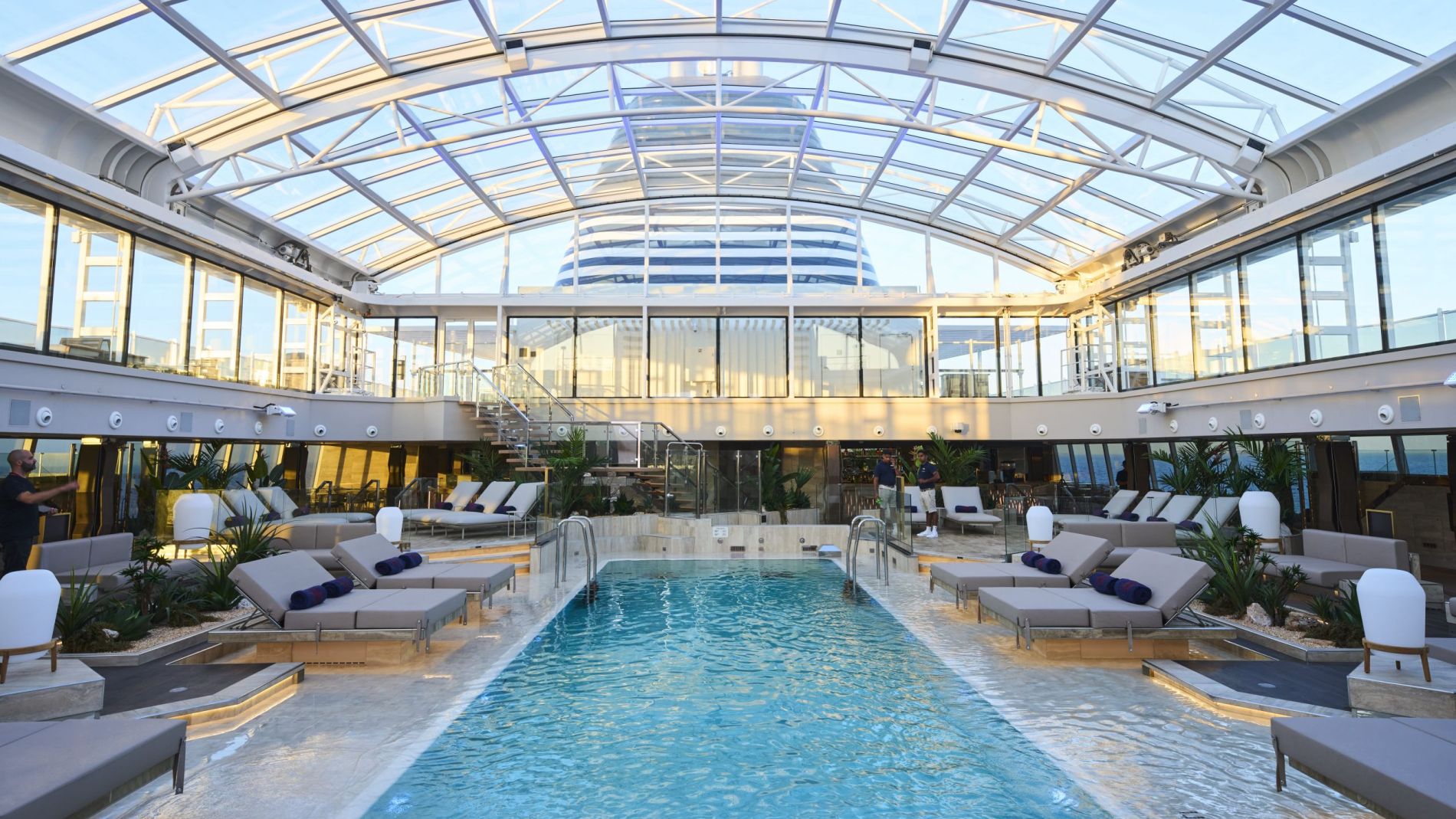 Innenpool in einem Glasdach-Atrium, umgeben von Liegen, Lounge-Sofas und Pflanzen auf einem Kreuzfahrtschiff.