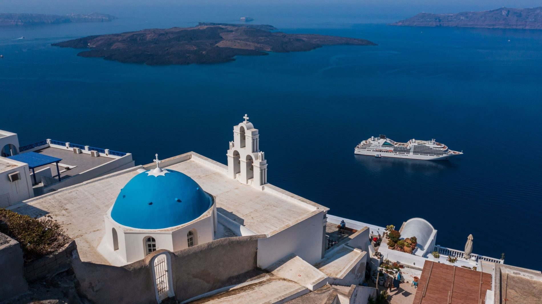 Weiße Kirche mit blauer Kuppel auf Santorini, Blick aufs Meer und Kreuzturm; Kreuzfahrtschiff im Hintergrund. Weiße Kirche mit blauer Kuppel auf Santorini, Blick aufs Meer und Kreuzturm; Kreuzfahrtschiff im Hintergrund.