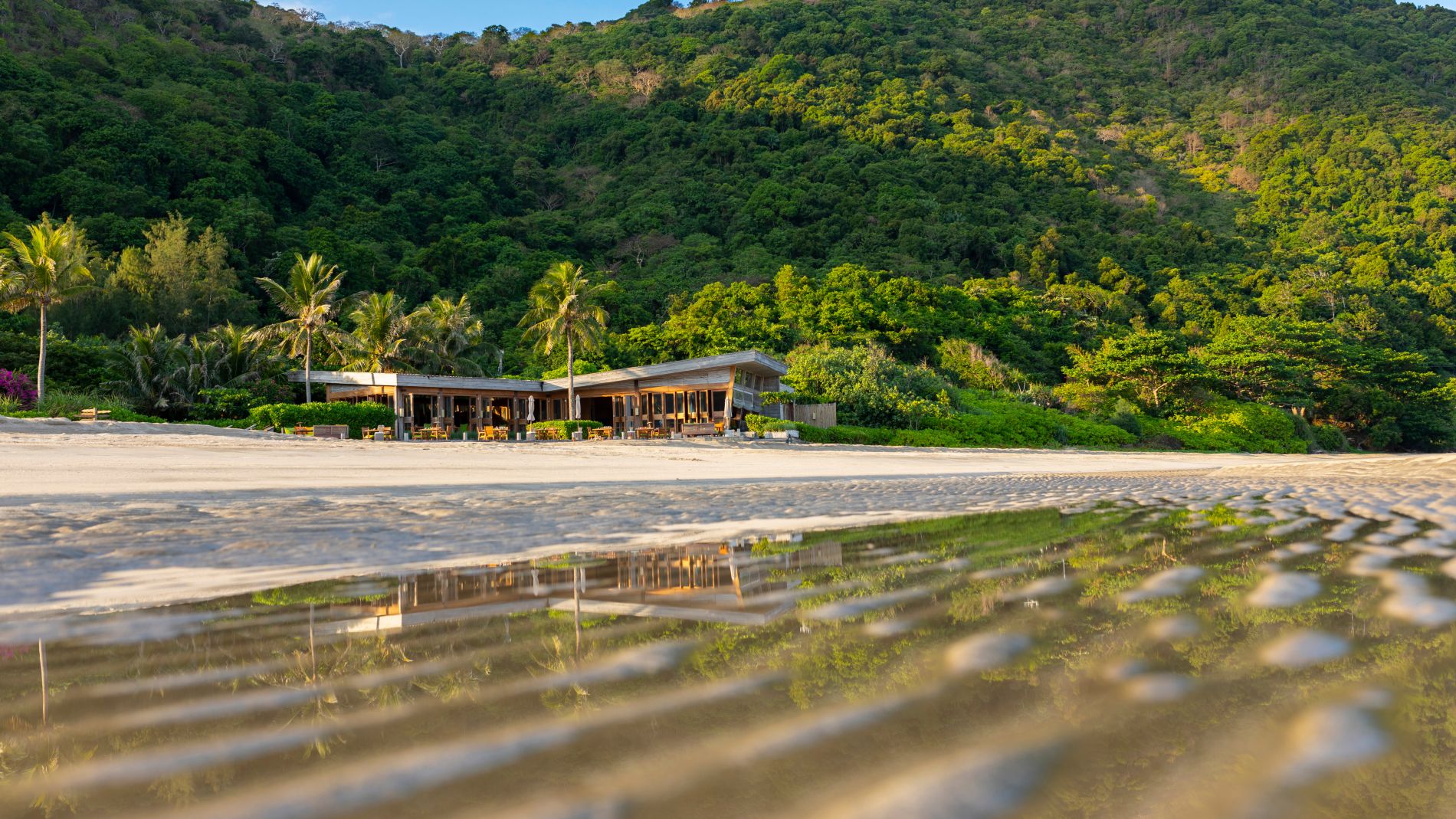 Strandrestaurant am Con Dao, hölzerner Bau mit Palmen, grüne Berge dahinter, Wasser reflektiert das Gebäude.