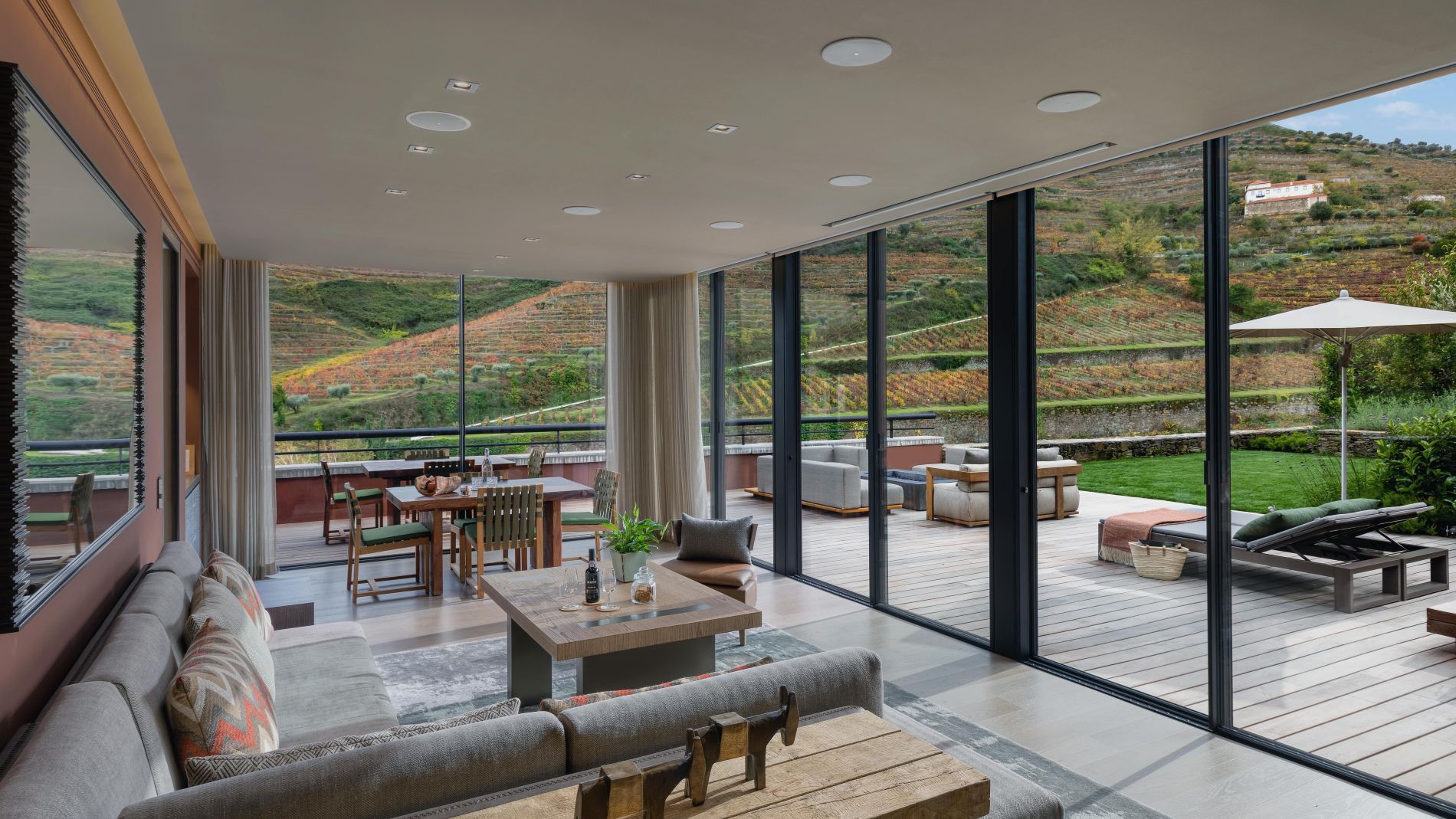 Wohnzimmer mit großen Glasfenstern, Blick auf terrassierte Weinberge; Innen- und Außenlounge mit Sofa und Esstisch.
