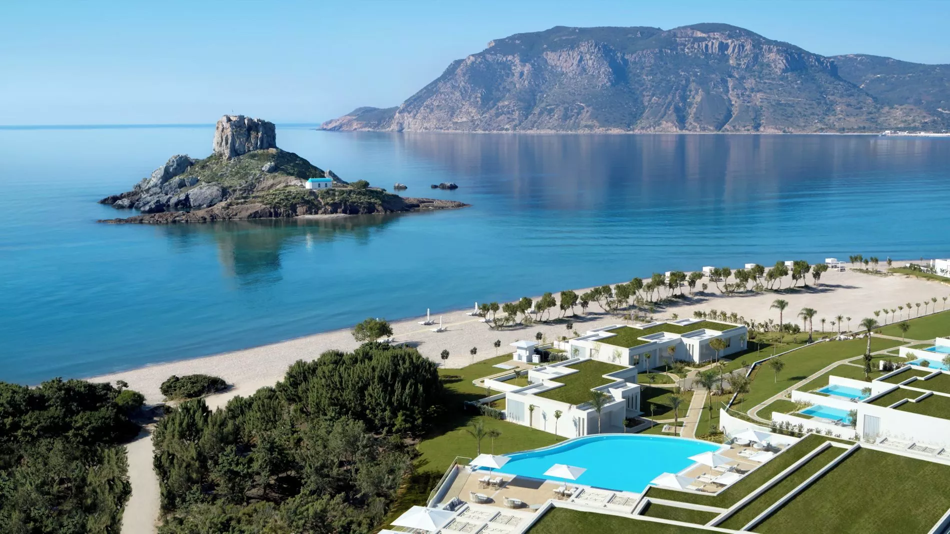 Luftaufnahme des Ikos Aria Resorts: Felsinsel, Strand, türkisblaues Meer und moderne Hotelanlage mit Poolbereich