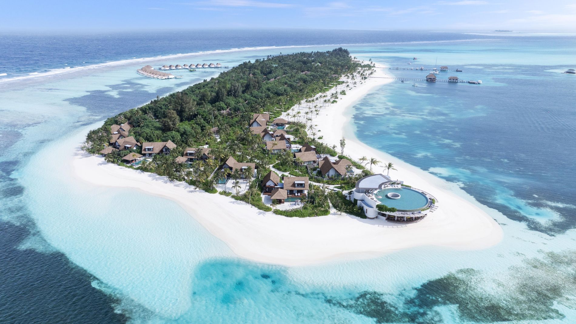 Luftaufnahme einer Inselresortanlage mit Palmen, braunen Bungalows und weißem Sandstrand, umgeben von türkisblauem Meer (Six Senses Kanuhura).