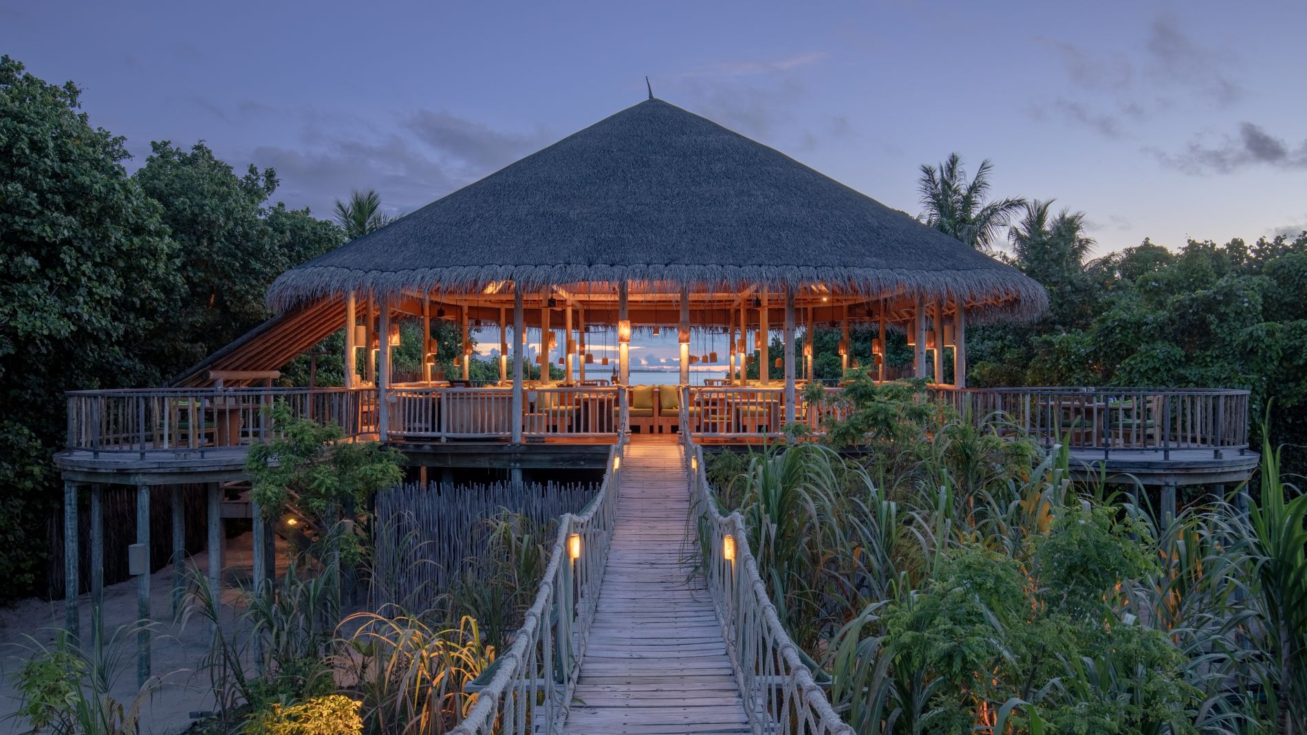 South Palm Resort Maldives, Six Senses: Strohdach-Pavillon auf Holzpfosten über Vegetation, beleuchtete Brücke zum Eingang.