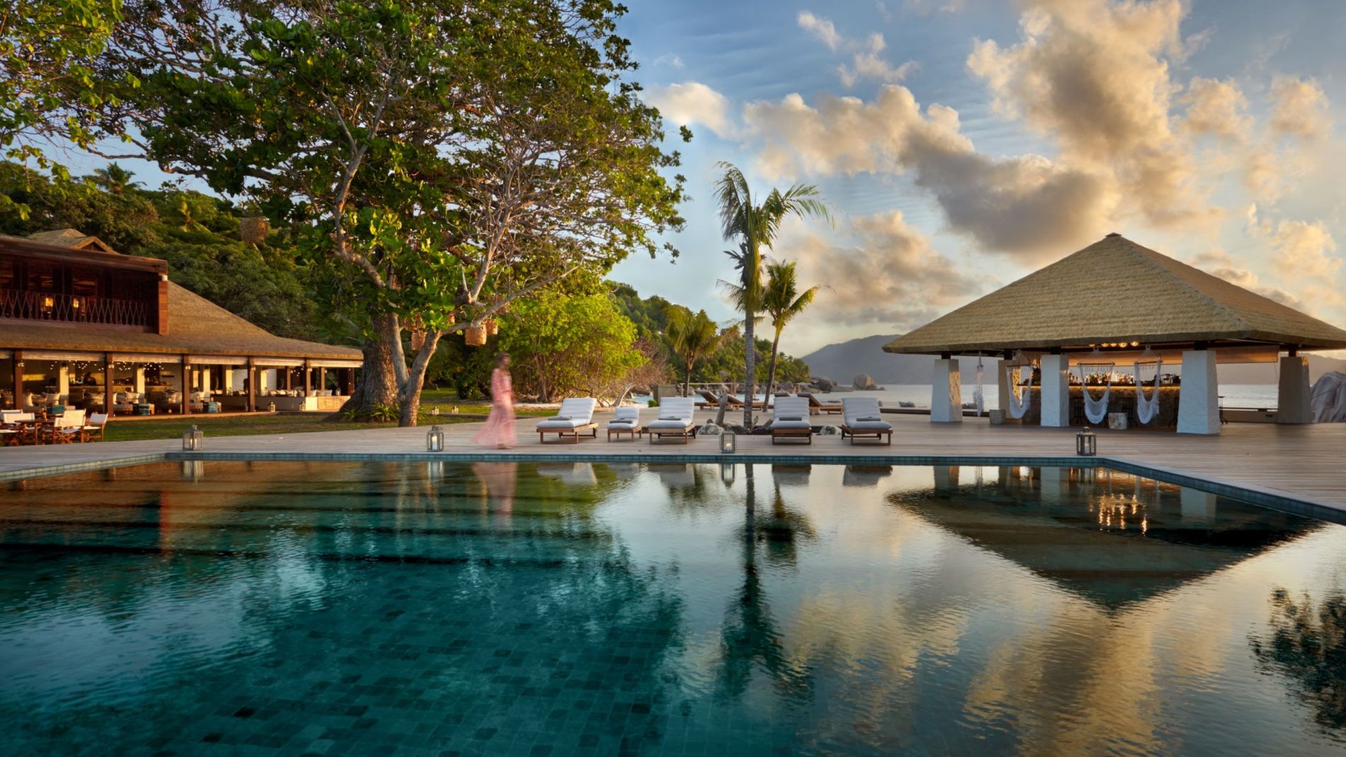 Six Senses Zil Pasyon: Poolbereich mit Palmen, Liegen und Pavillon am Strand; Wasser reflektiert Abendhimmel.