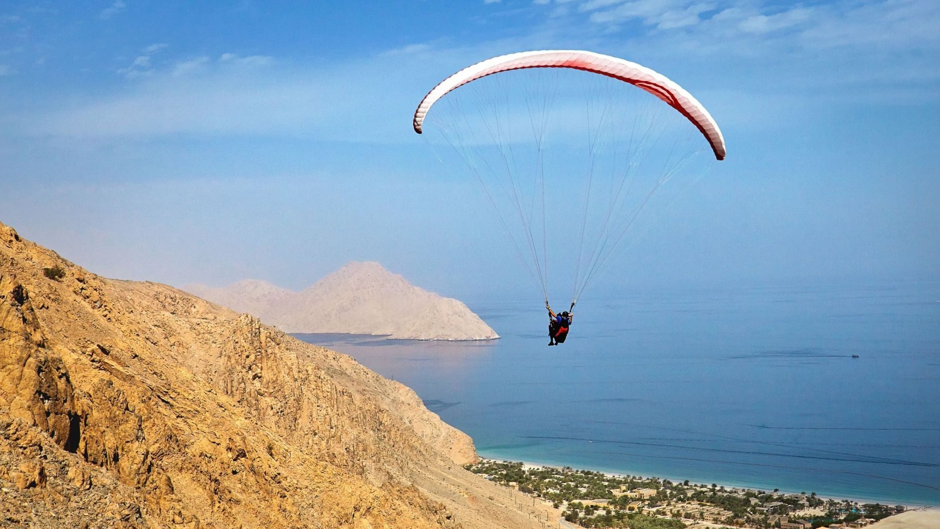Paragleiter mit weißem Schirm über dem Meer vor Zighy Bay; felsige Küste links, Strand unten.