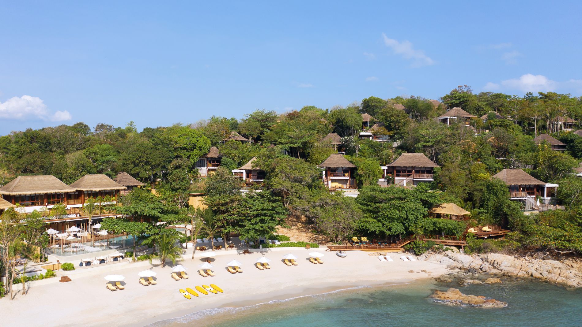 Luftaufnahme des SS Samui Resorts: Strand mit Liegen und gelben Kajaks, dahinter Hüttenvillen im Strandwald.