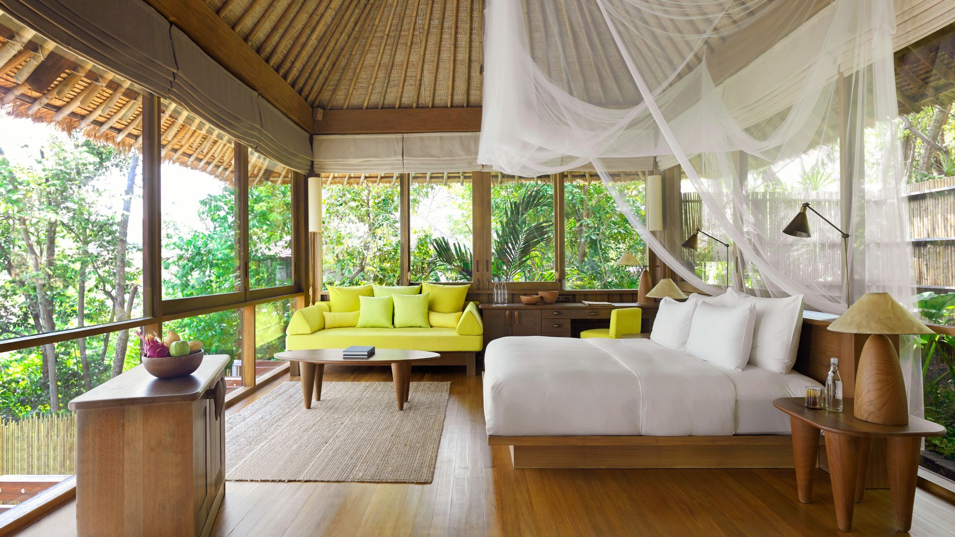 Interieur der SS Samui Hideaway Pool Villa: großes Bett mit Moskitonetz, gelbes Sofa, Holzmoebel, breite Fenster zum Garten