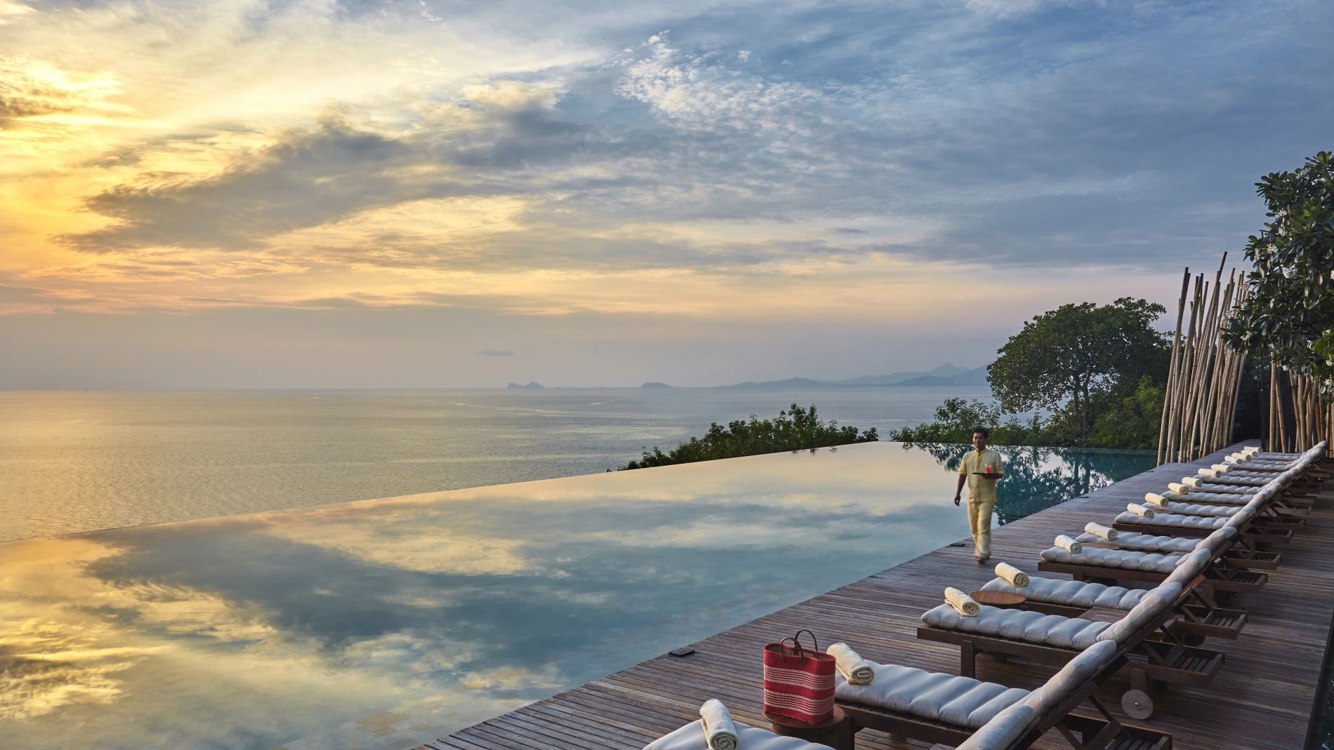 Infinity-Pool mit Meerblick und Sonnenuntergang, Holzdeck mit Liegen und Tüchern, Mitarbeiter mit Servierkorb geht vorbei.