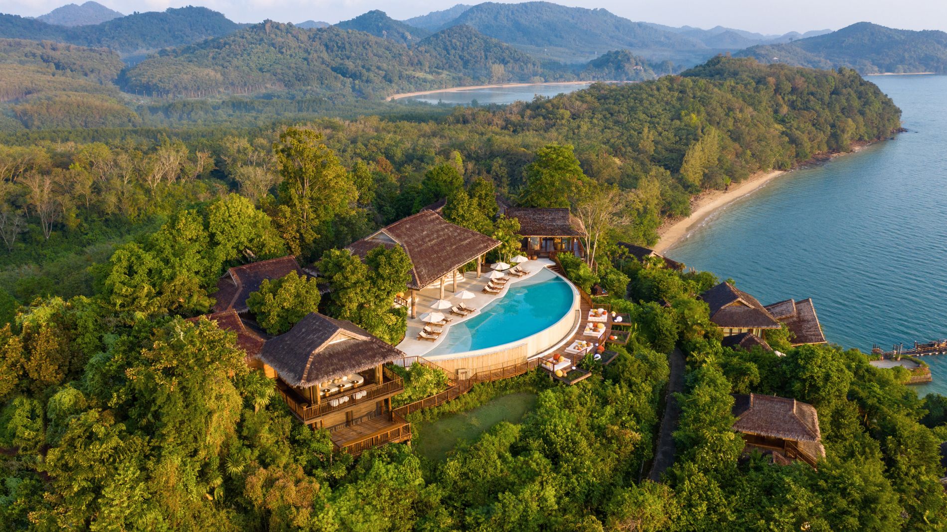 Luftaufnahme eines Hangresorts mit rundem Pool, Strohdach-Villen, Strandzugang und Wald am Meer