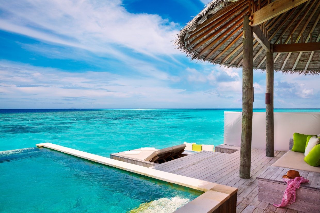 Overwater-Villa mit Pooldeck am türkisblauen Meer; Strohdach, Holzdeck, Loungebereich mit grünen Kissen.