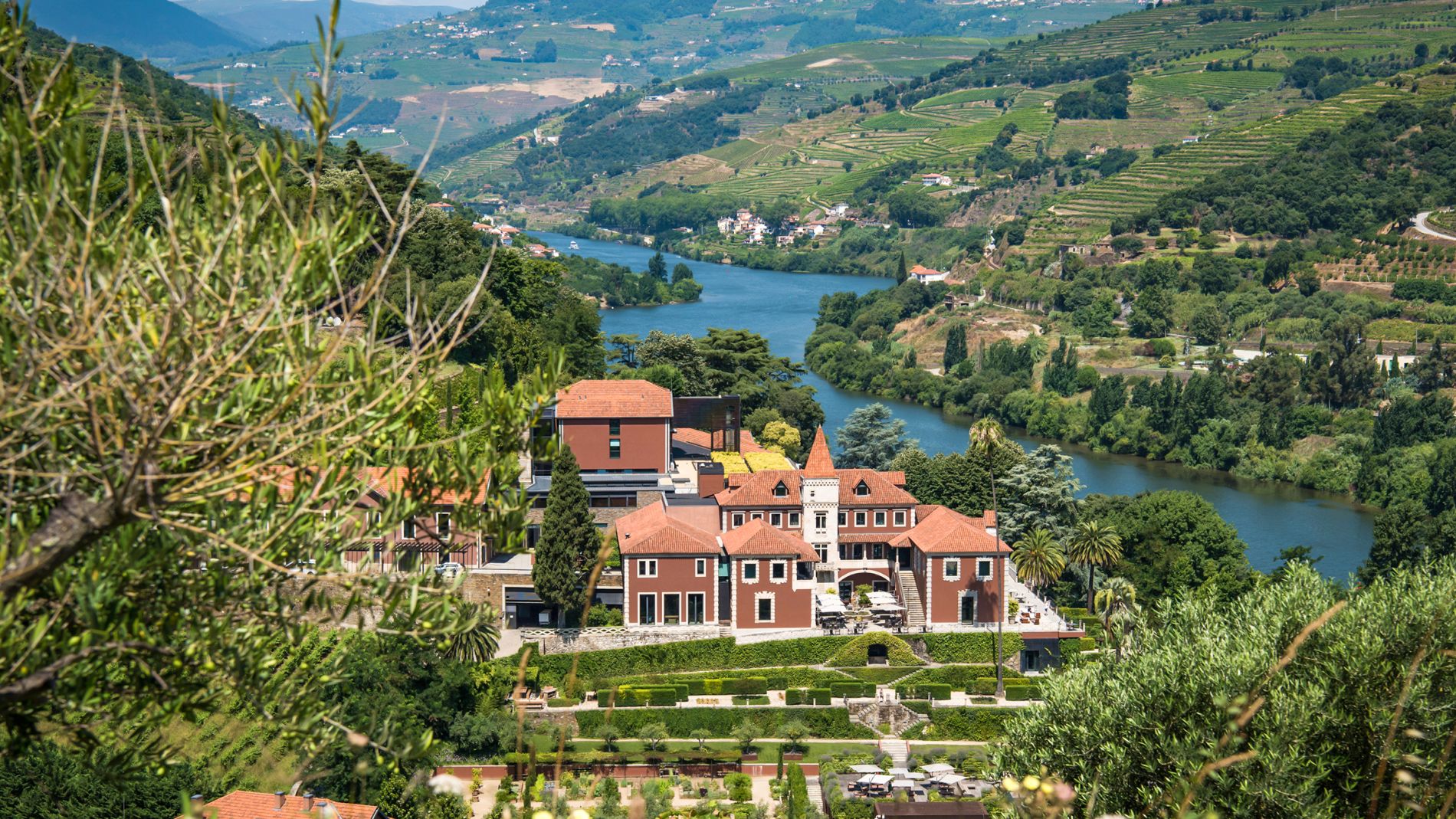 Blick über das Douro-Tal auf Terrassenfelder, Fluss und ein rotes Villengebäude im Vordergrund.