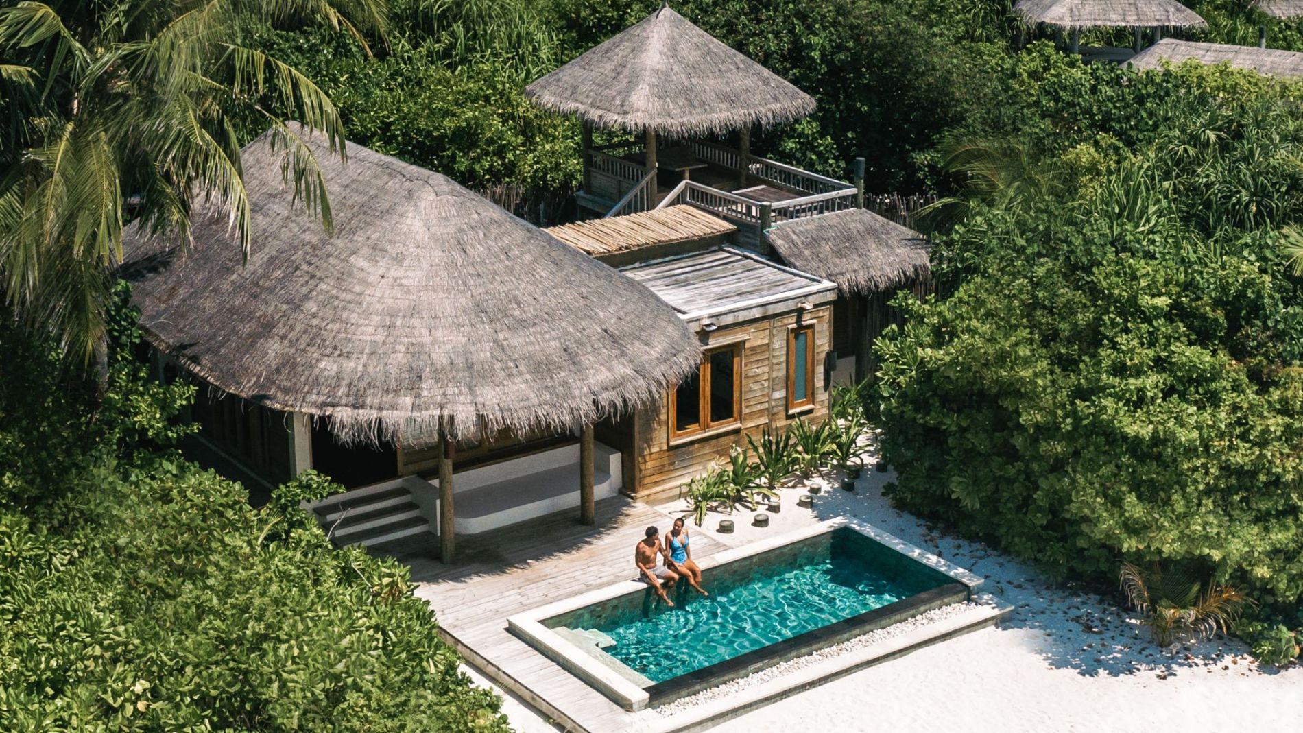 Luftaufnahme einer Beach-Villa mit Strohdach, kleinem Pool, Palmen und Sand; turquoise Lagune und Overwater-Bungalows im Hintergrund