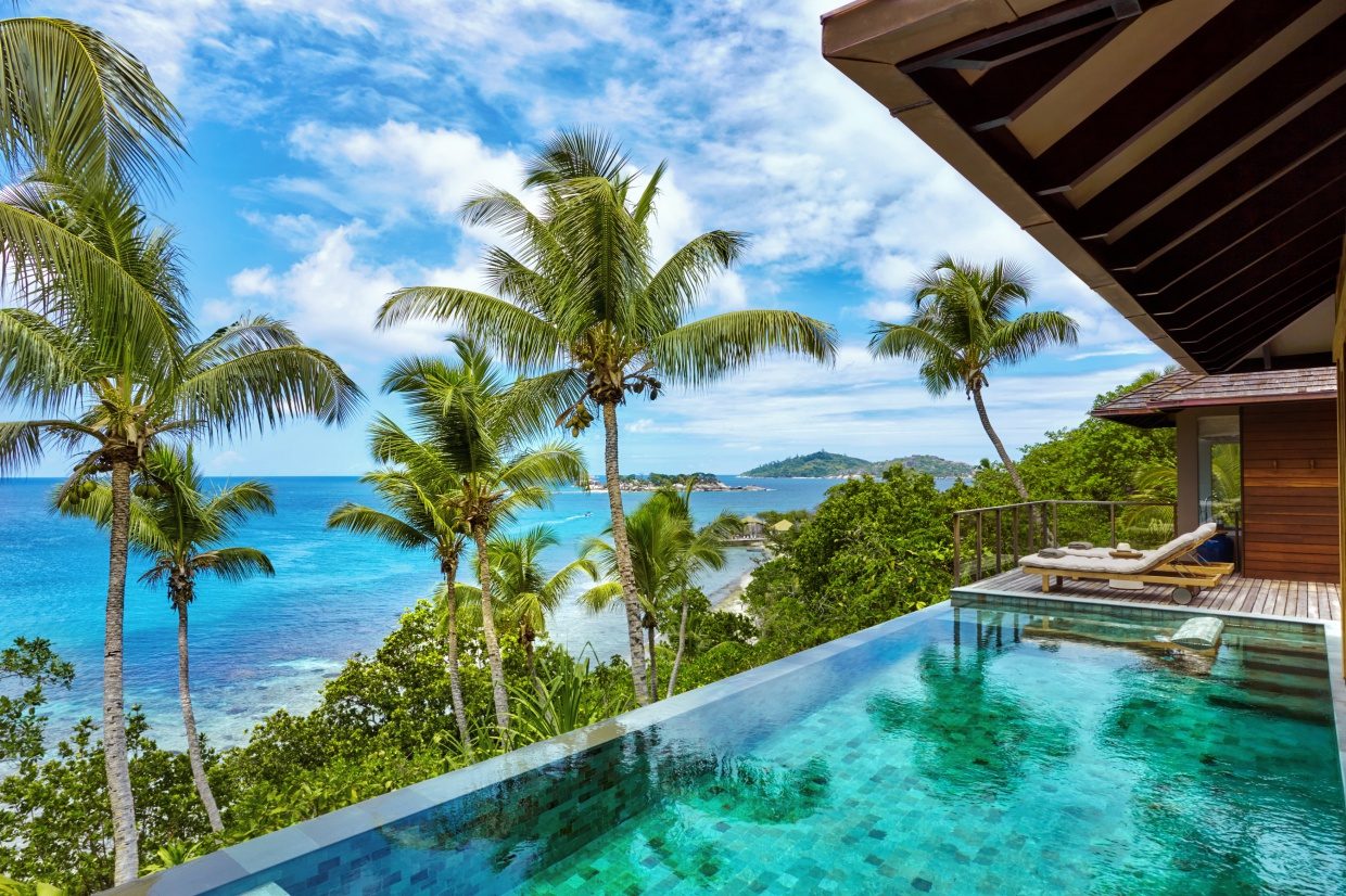 Infinity-Pool mit Meerblick, Palmen und tropischer Vegetation; Holzdeck mit Liege vor einer Villa.