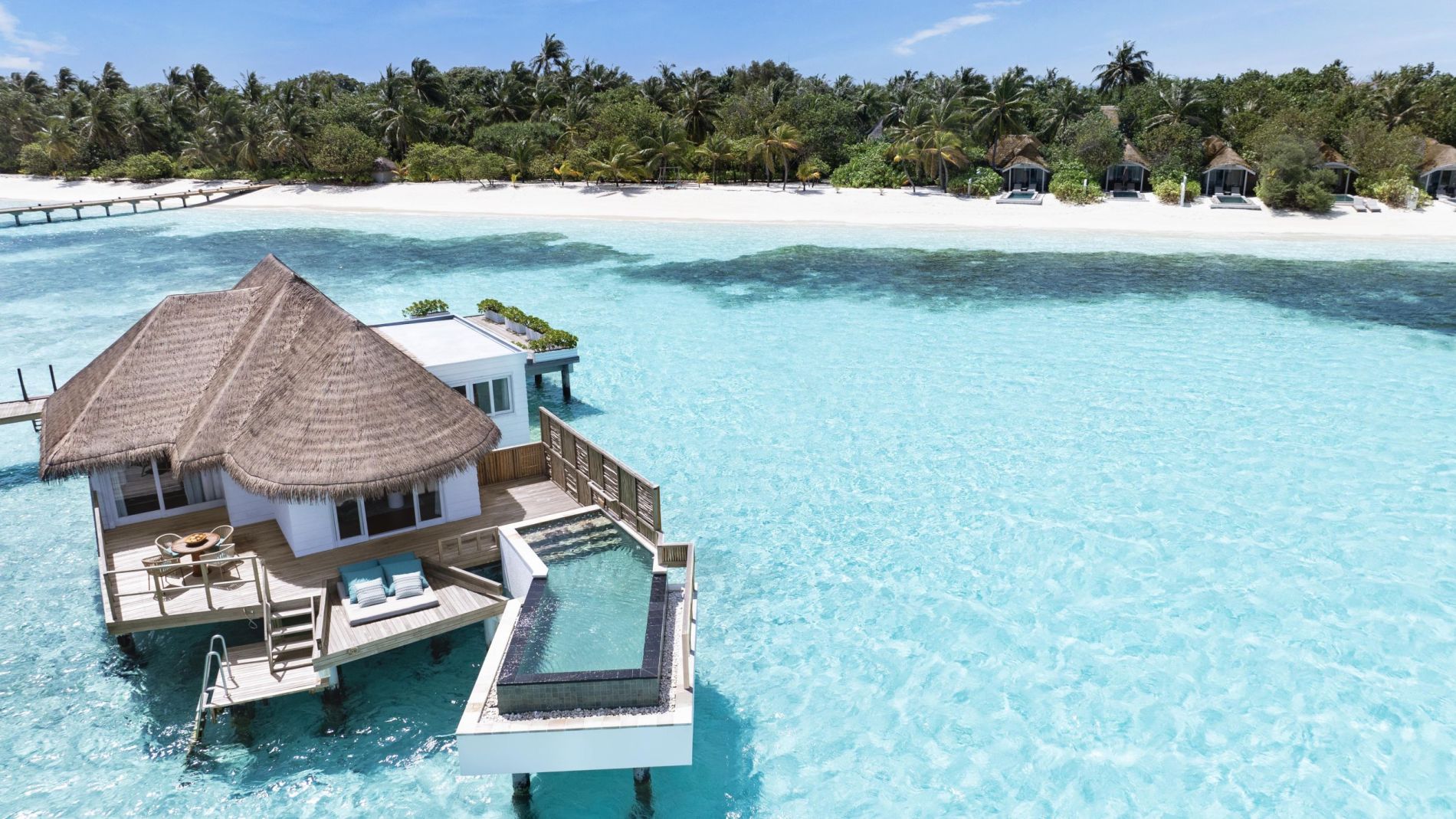 Überwasser-Bungalow mit Strohdach auf türkisblauem Meer, Holzdeck mit Sitzbereich und kleinem Becken, Strand und Palmen im Hintergrund