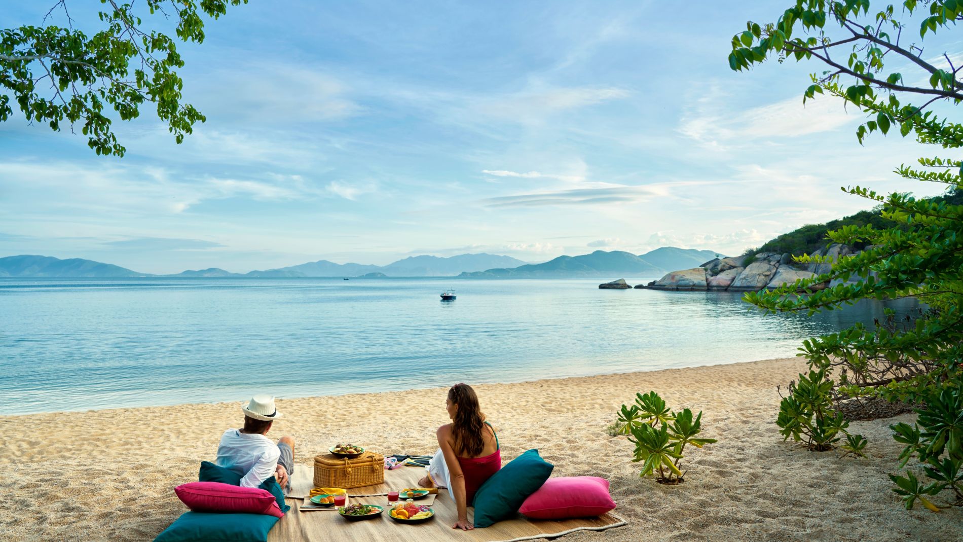 Paar genießt Picknick am Sandstrand, Korb und Speisen auf Decke, ruhiges Meer, grüne Büsche am Ufer, Inseln am Horizont.