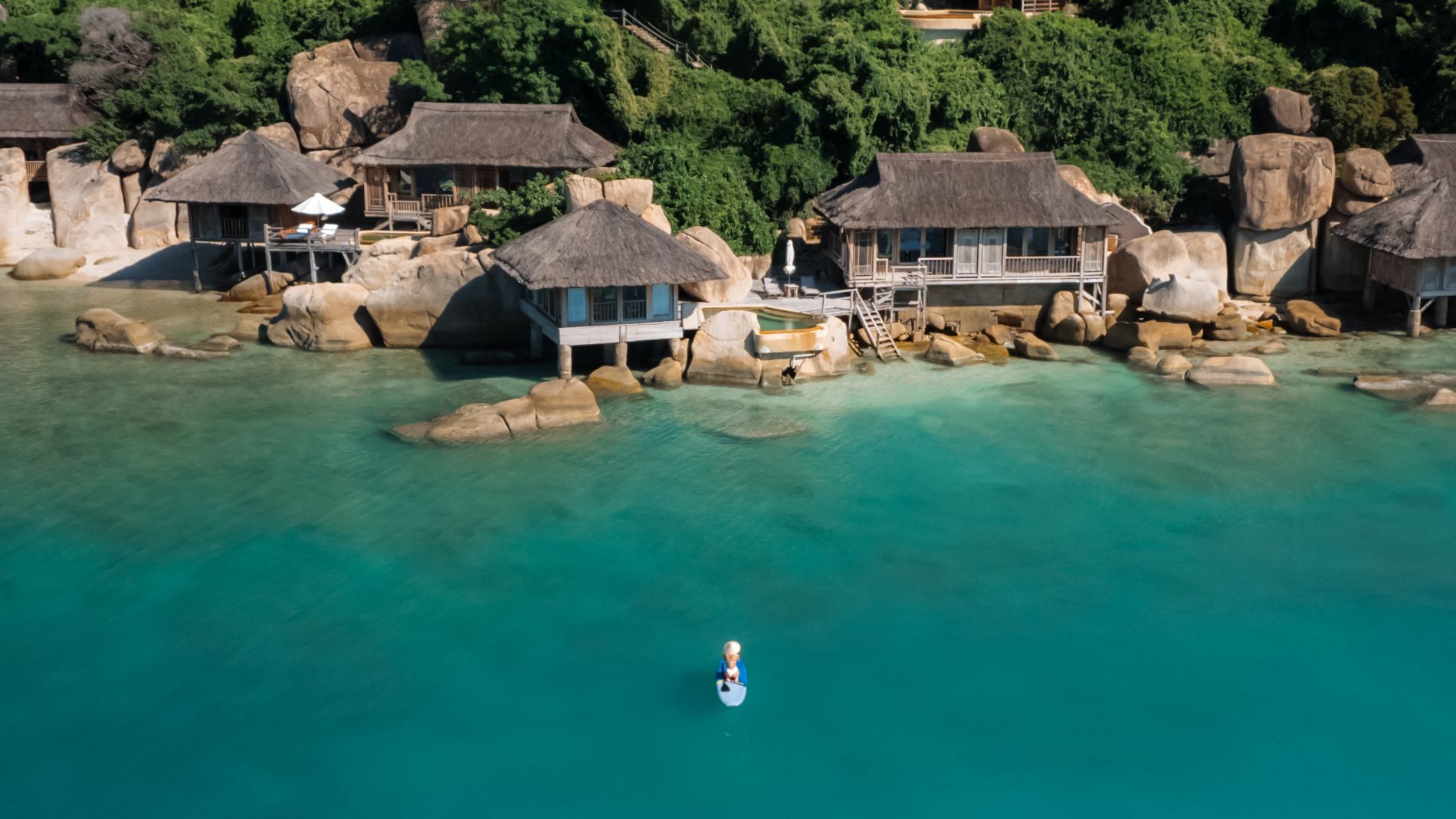 Überblick über eine Küstenresort-Siedlung mit Strohdach-Häusern an Felsen, grünem Hang und turquoise Wasser, Person auf Stand-Up-Paddleboard.