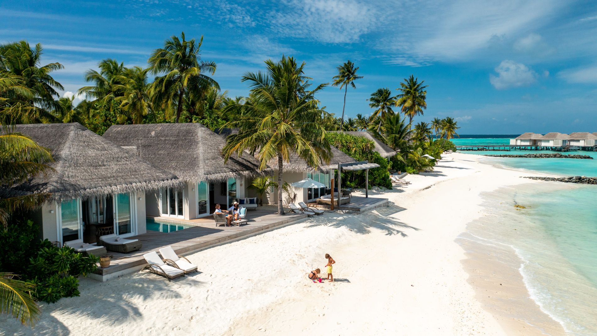 Weißer Sandstrand, Palmen, Reetdach-Villen am Wasser, Liegen auf Deck, türkisblaues Meer im Hintergrund (Baglioni Maldives)