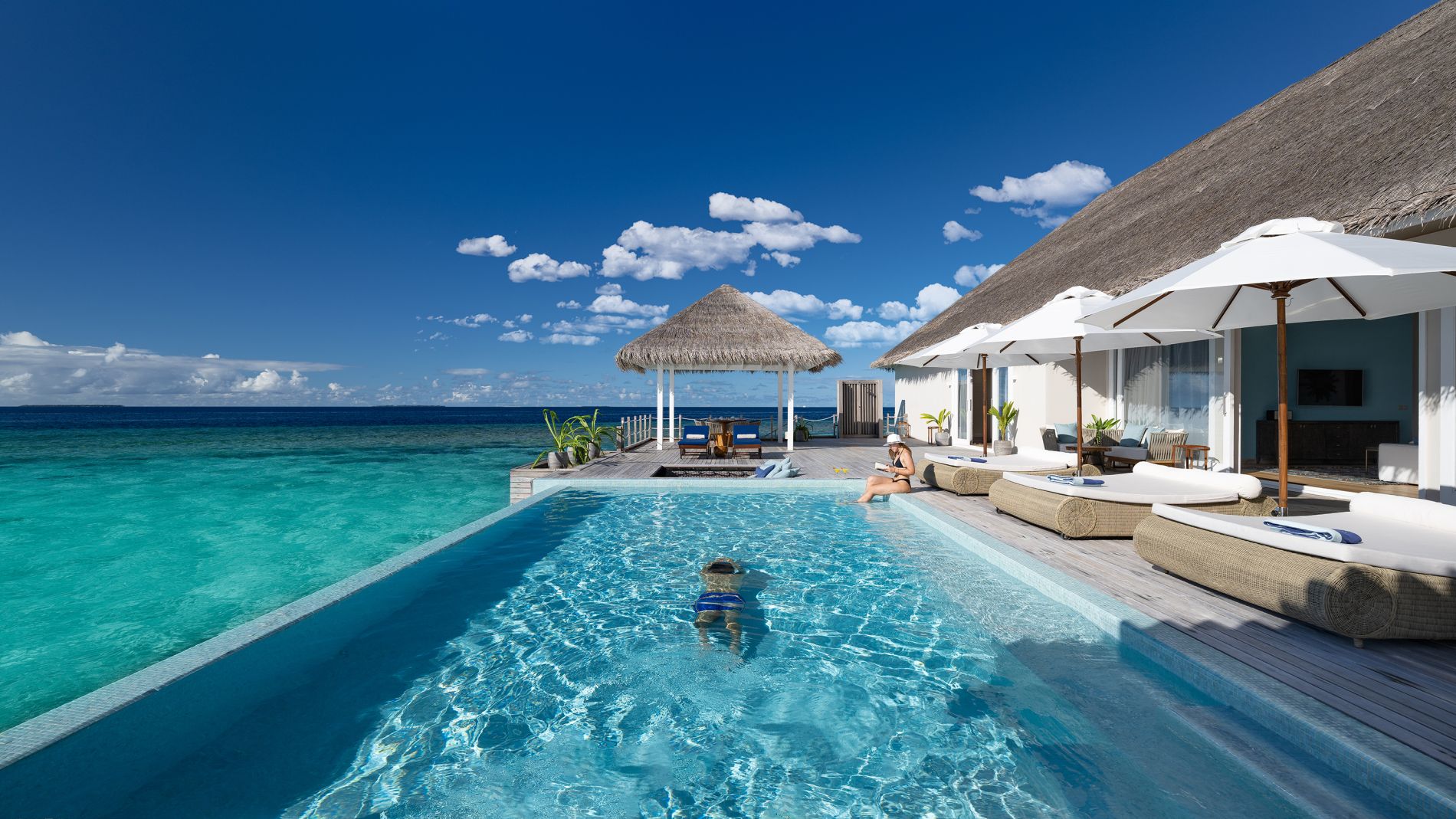Infinity-Pool am Baglioni Resort Maldives, türkisblaues Meer, Reetdachvillen, loungem Liegen, Sonnenschirme; eine Person schwimmt im Wasser