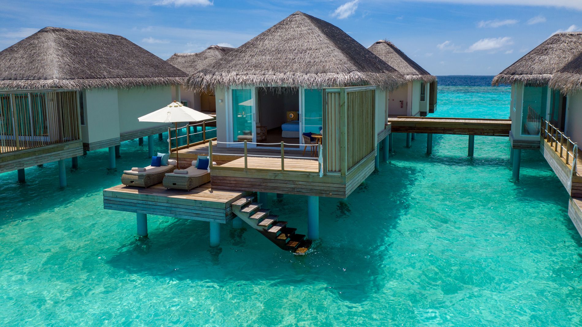 Überwasser-Villen mit Strohdächern, Holzdecks über türkisblauem Meer; Loungebereich und Stufen ins Wasser.