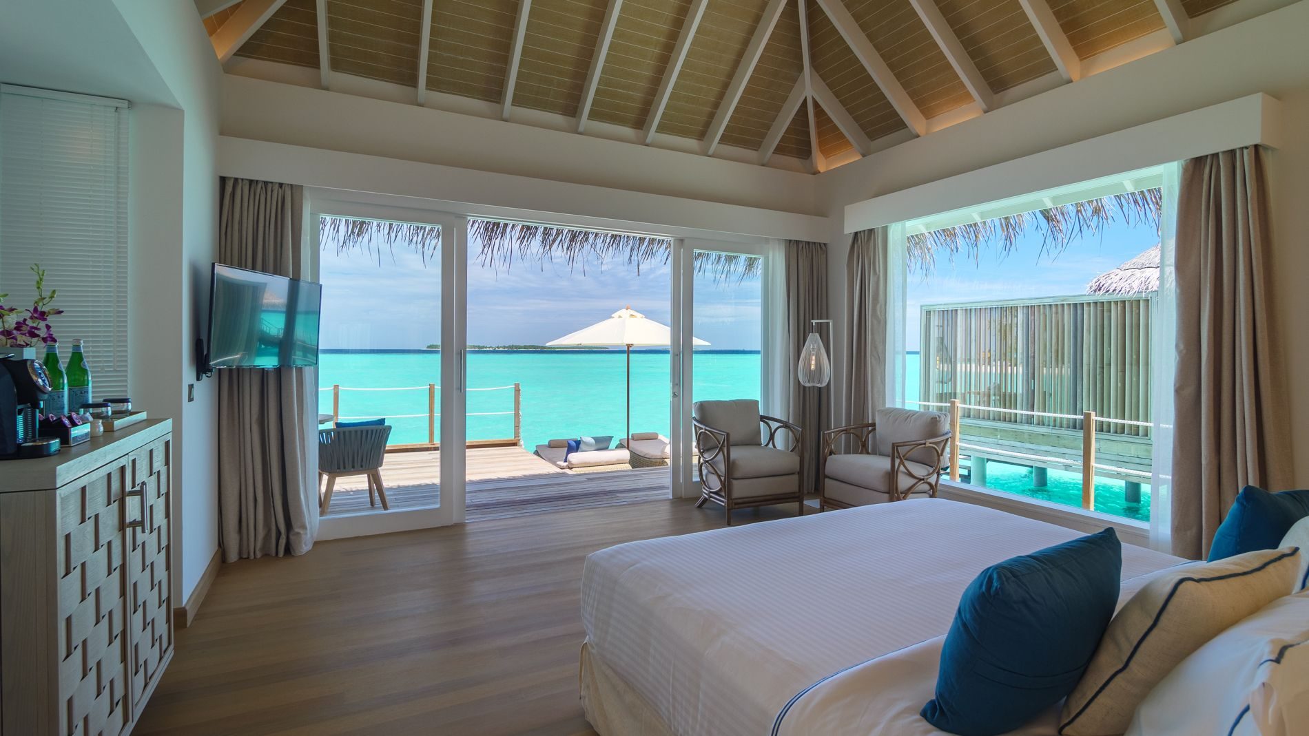 Bettzimmer einer Wasser-Villa im Baglioni Resort Maldives; Blick auf türkisblaues Meer, Deck, Lounge-Sessel, große Fenster.
