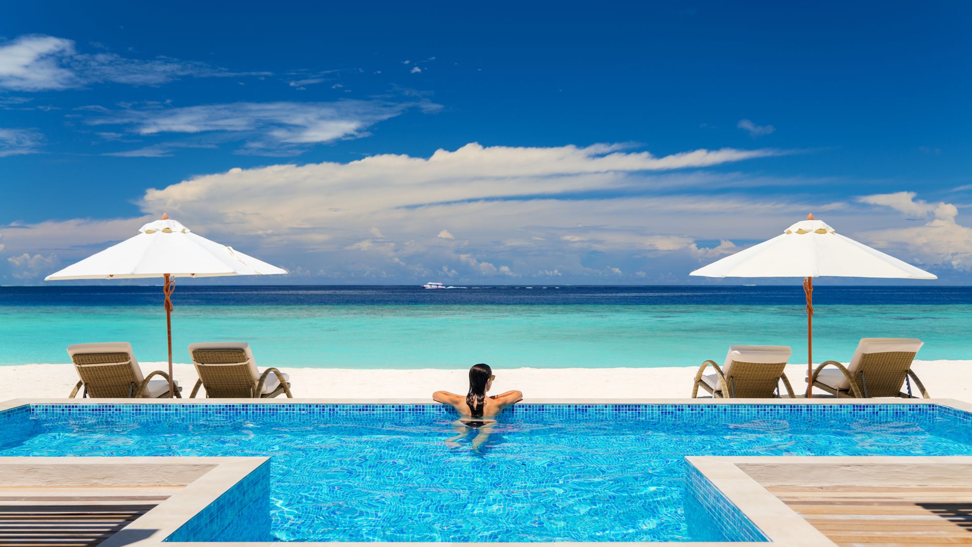 Person sitzt im Pool und blickt aufs Meer; zwei Liegen mit weißen Sonnenschirmen am Strand, türkisblaues Wasser, Baglioni Resort Maldives