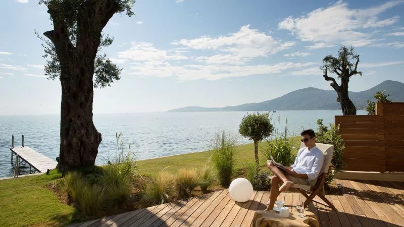 Mann in weißem Hemd sitzt lesend auf Deckstuhl am Ufersee, Blick aufs Meer, Olivenbäume und Berglandschaft.