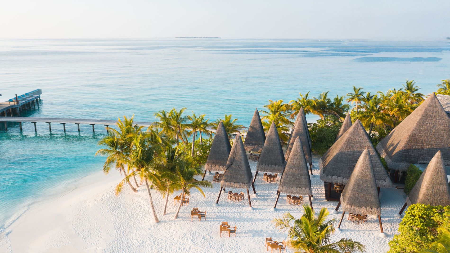 Luftaufnahme eines türkisfarbenen Strandabschnitts mit Palmen, Strohut-Hütten, Tischen und Liegen; Pier im Hintergrund