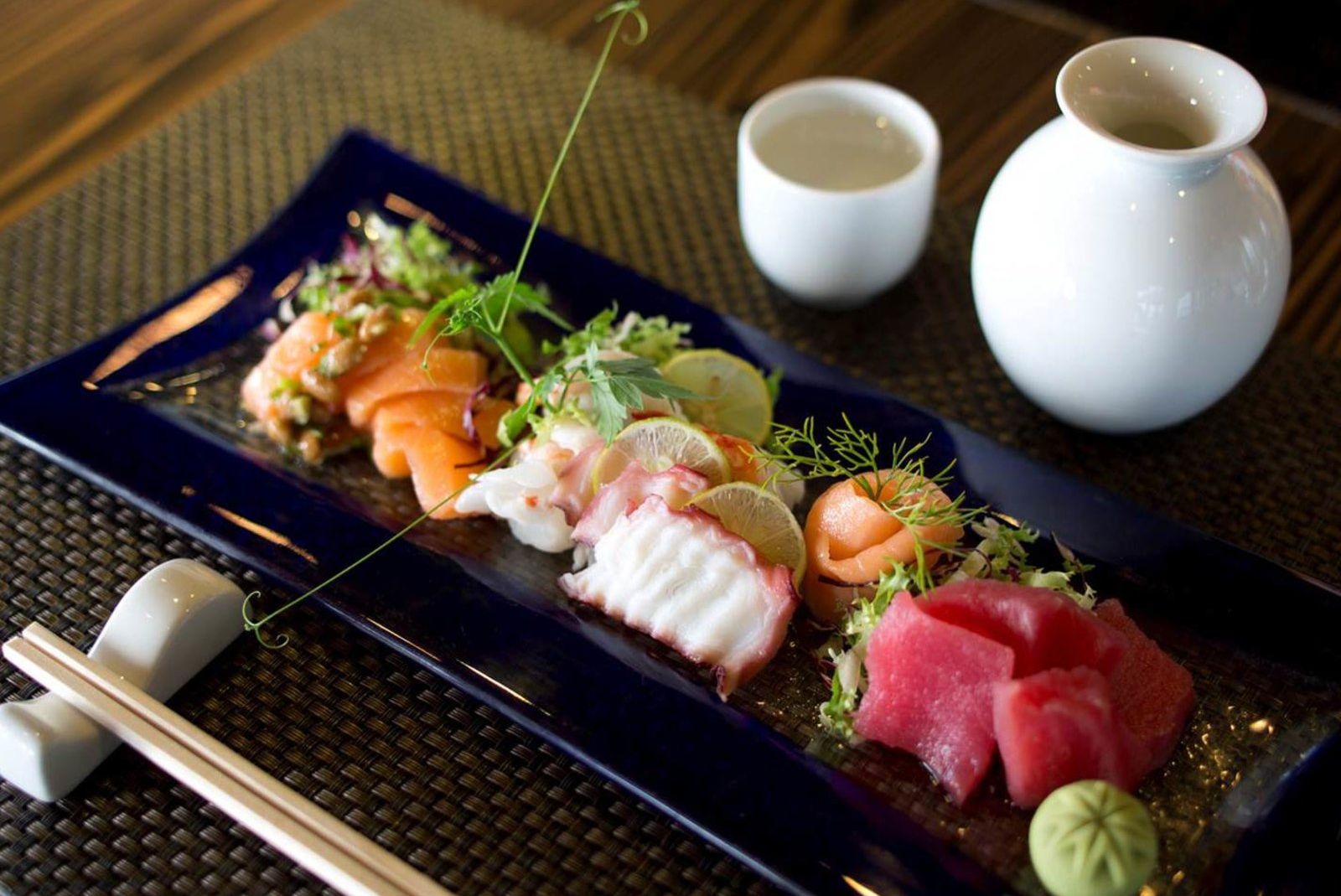 Sushi-Platte mit verschiedenen Rahmungen, Wasabi, Sojasauce und einem weißen Sake-Behälter auf einem Tisch.