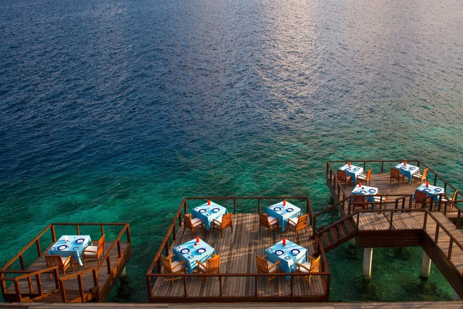 Holzplattformen mit Tischen und Stühlen am Meer, türkisfarbenes Wasser im Hintergrund.