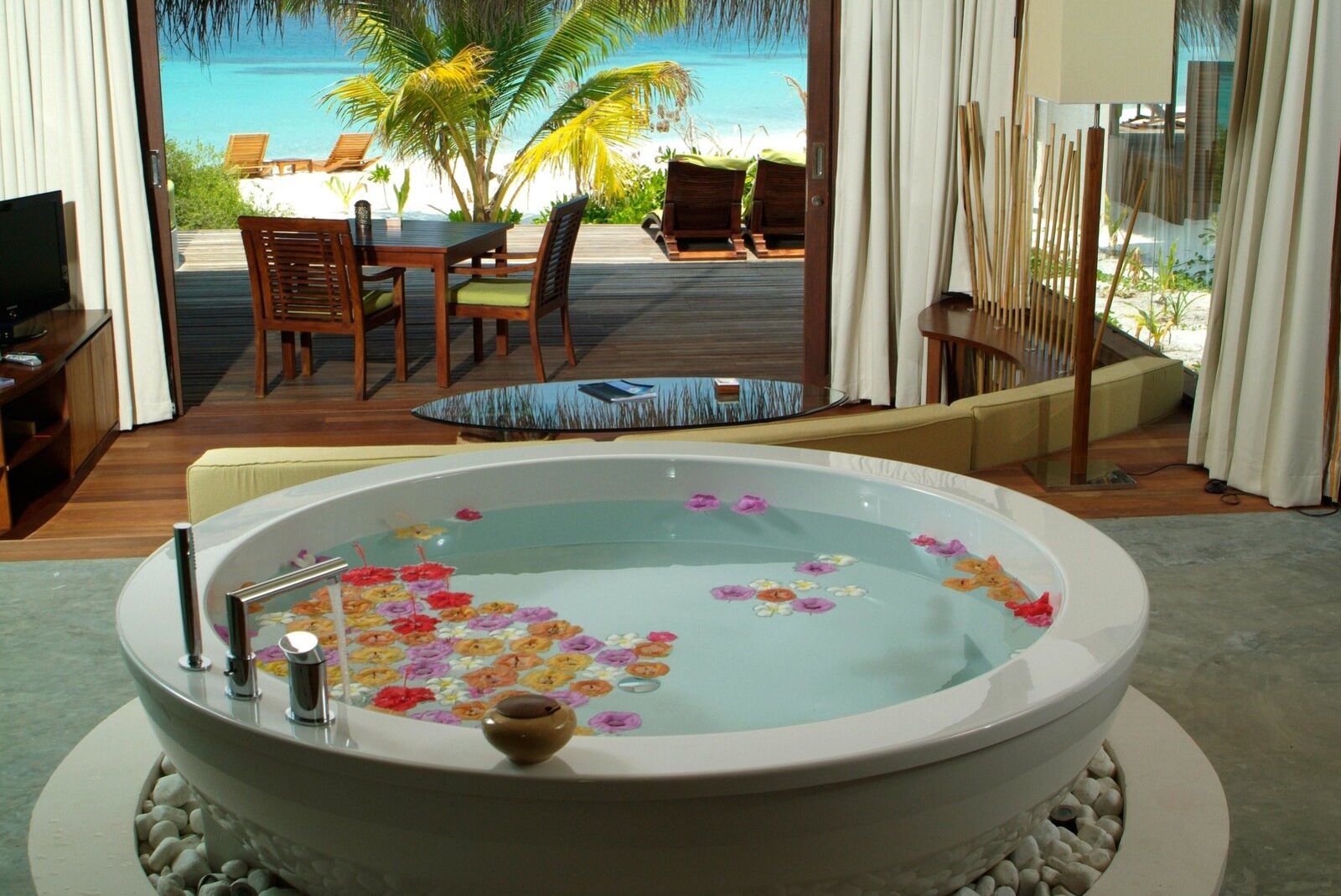 Blick auf ein Zimmer mit Badewanne, gefüllt mit Blüten, Blick auf Terrasse und Meer im Hintergrund.