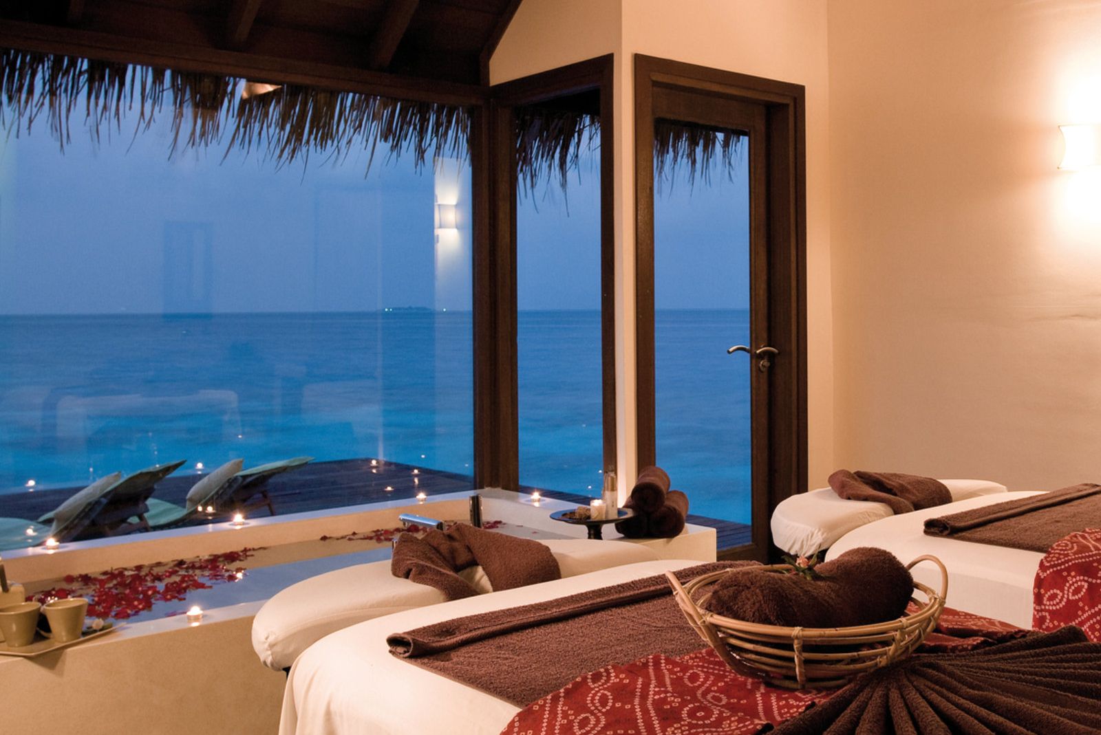 Wellnessbereich mit zwei Behandlungsliegen, Blick auf das Meer, dekoriert mit Kerzen und Blumenblättern.