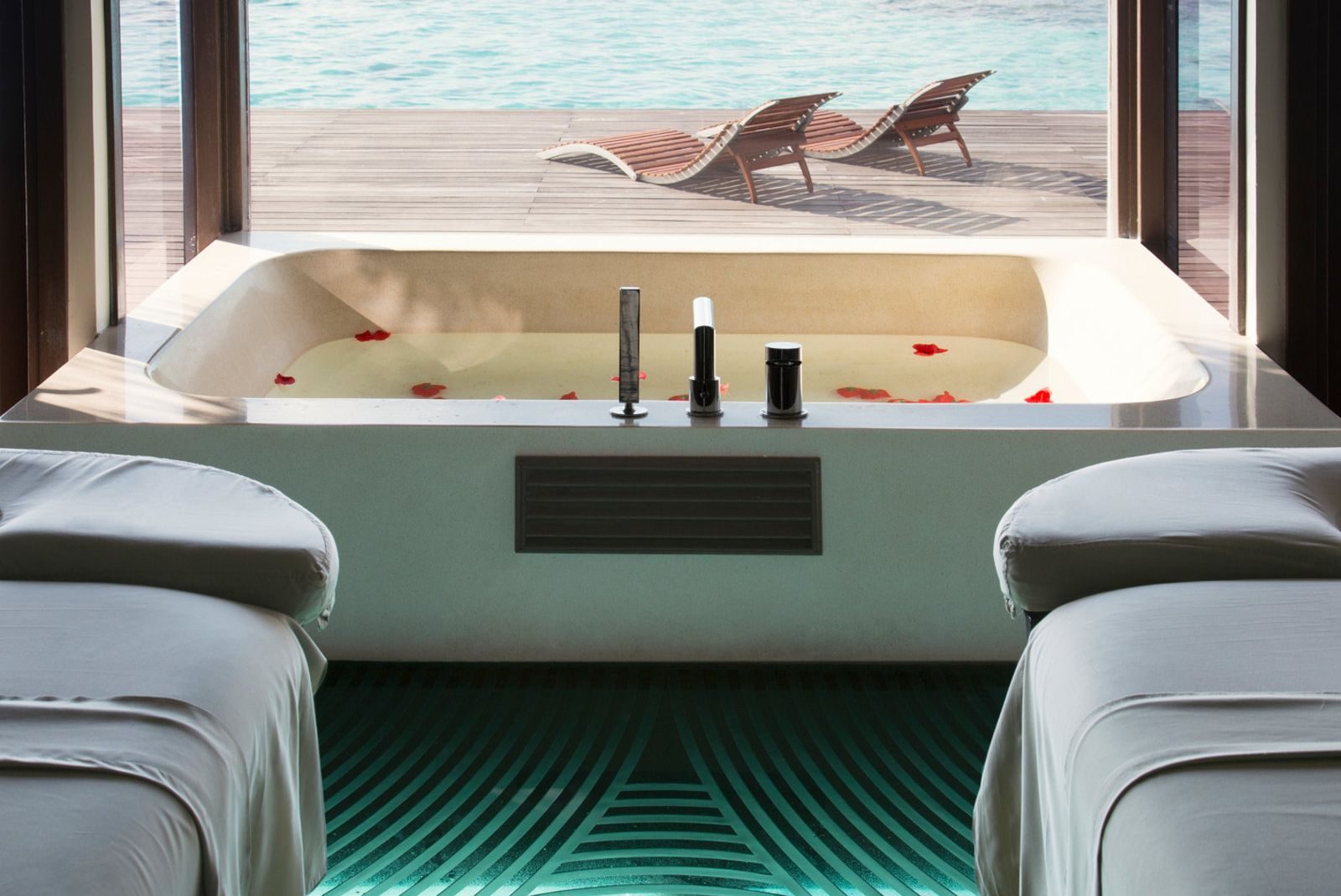 Blick auf ein Spa mit einem Wannenbereich, umgeben von zwei Liegen und Blick auf das Meer. Roseblätter schwimmen im Wasser.