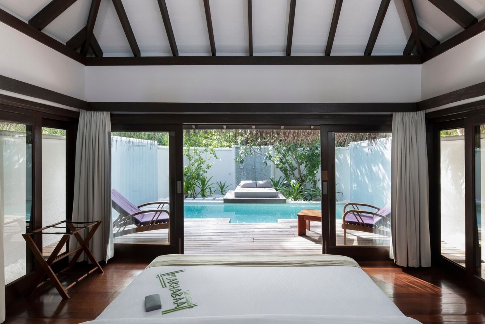 Blick auf ein modernes Zimmer mit Bett, verglaster Wand, Pool und Außenmöbeln im Hintergrund.