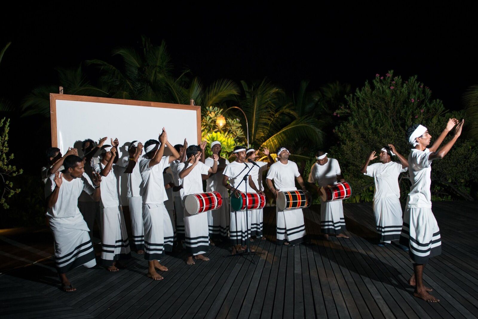 Tanzende Musiker in traditionellen weißen Kleidern mit Trommeln auf einem Holzdeck bei Nacht.