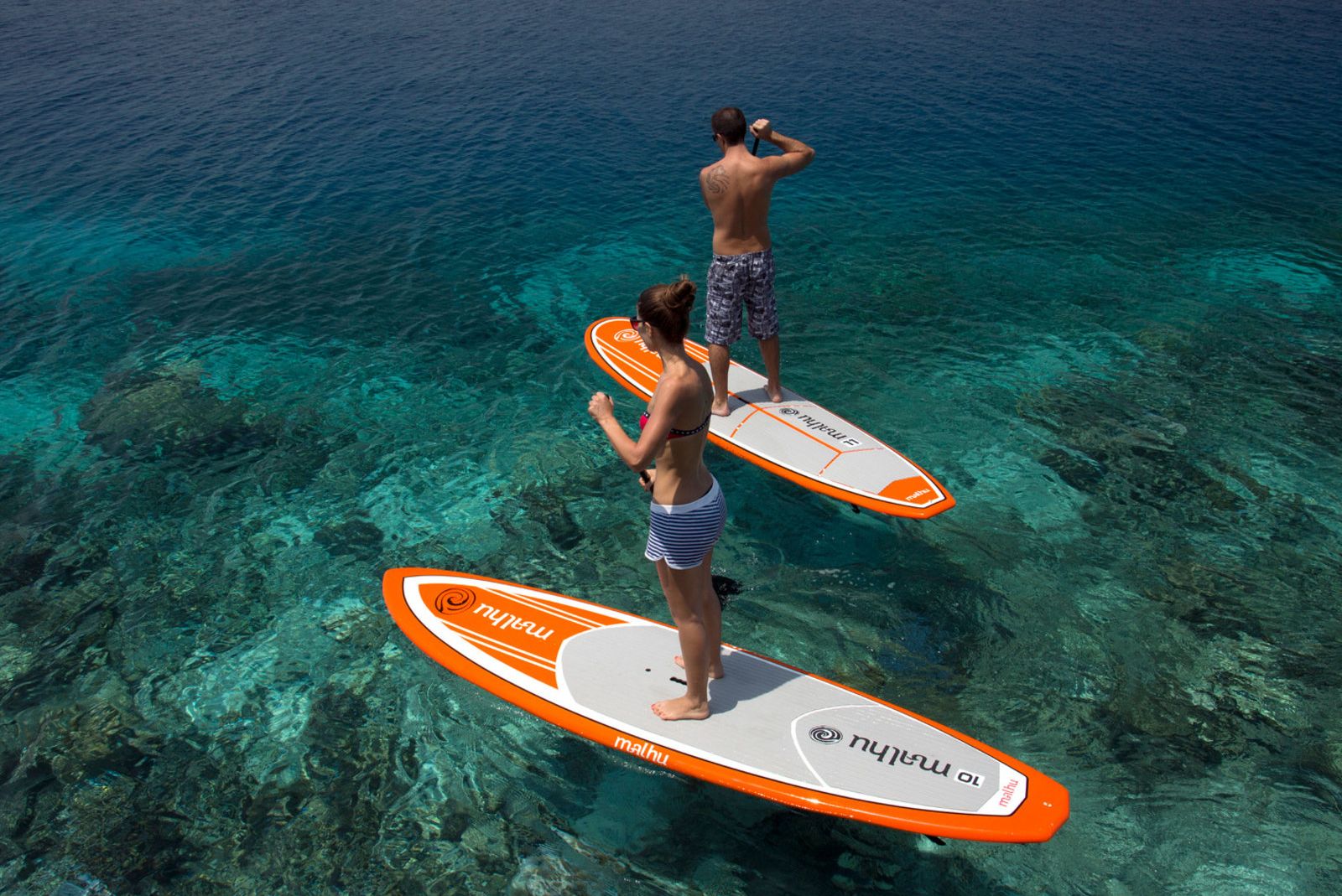 Zwei Personen stehen auf Stand-Up-Paddle-Boards über klarem, türkisfarbenem Wasser.