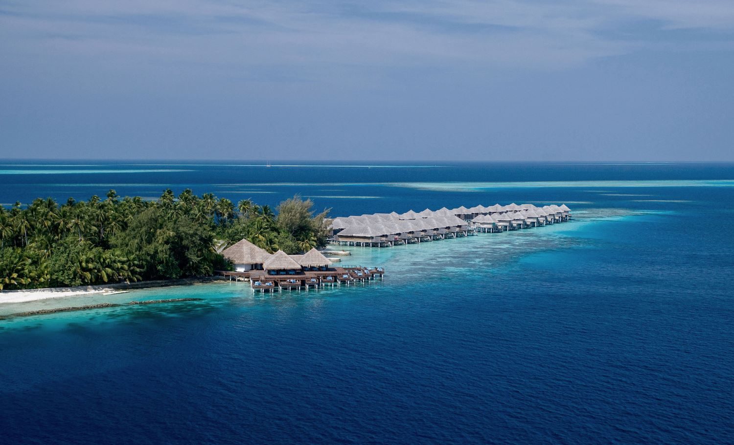 Luftaufnahme eines Resorts mit Überwasserbungalows und tropischen Pflanzen, umgeben von blauem Wasser und Korallenriffen.