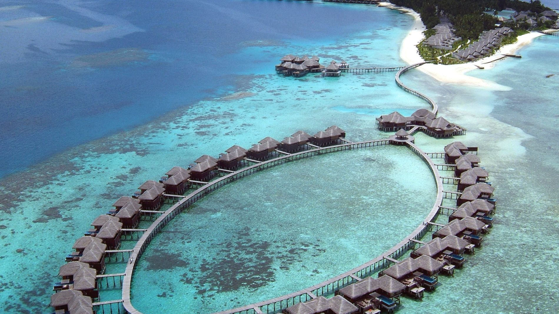 Luftaufnahme eines Resorts mit über dem Wasserstehenden Bungalows und klarem, blauem Wasser umgeben von Korallenriffen.