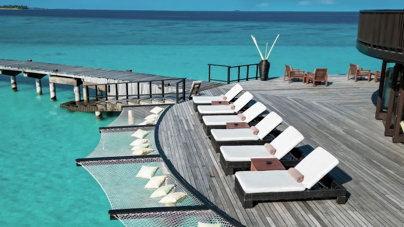 Liegemöbel und Kissen auf einem Holzdeck über kristallklarem Wasser mit Himmel und Wolken im Hintergrund.