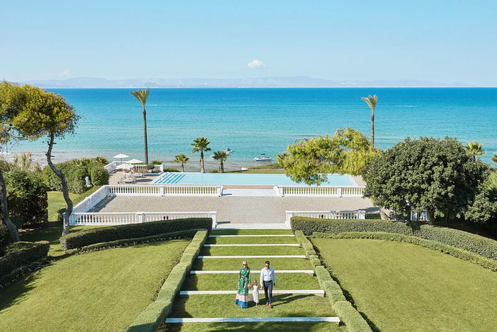 Paar geht einen Gartenweg hinunter, im Hintergrund das Meer, Palmen und ein Schwimmbad sichtbar.