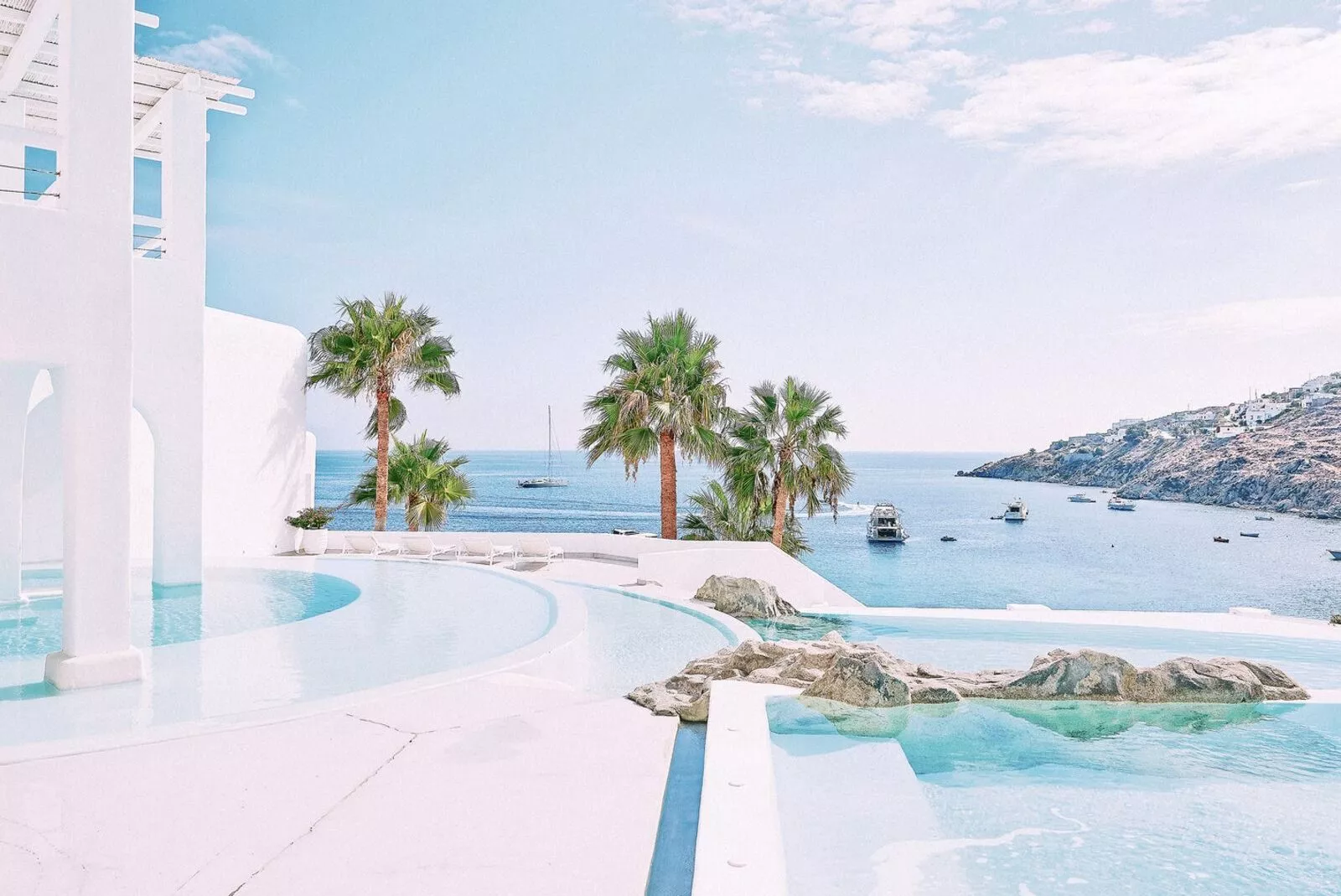 Poollandschaft mit Palmen, Blick auf das Meer und Boote, helles blaues Himmel, moderne Architektur in Mykonos.