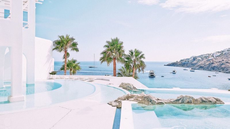 Poollandschaft mit Palmen, Blick auf das Meer und Boote, helles blaues Himmel, moderne Architektur in Mykonos.