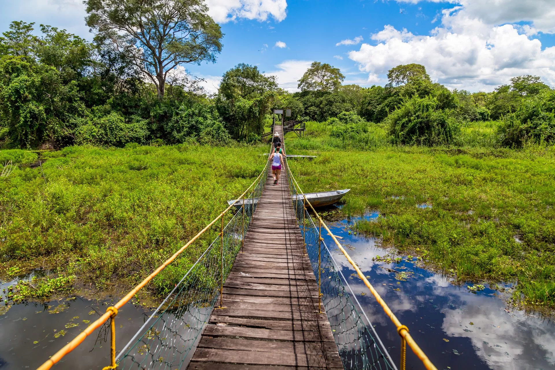 Zwei Personen gehen über eine hölzerne Hängebrücke in einer grünen Landschaft des Pantanal in Brasilien, umgeben von Wasser und üppiger Vegetation.